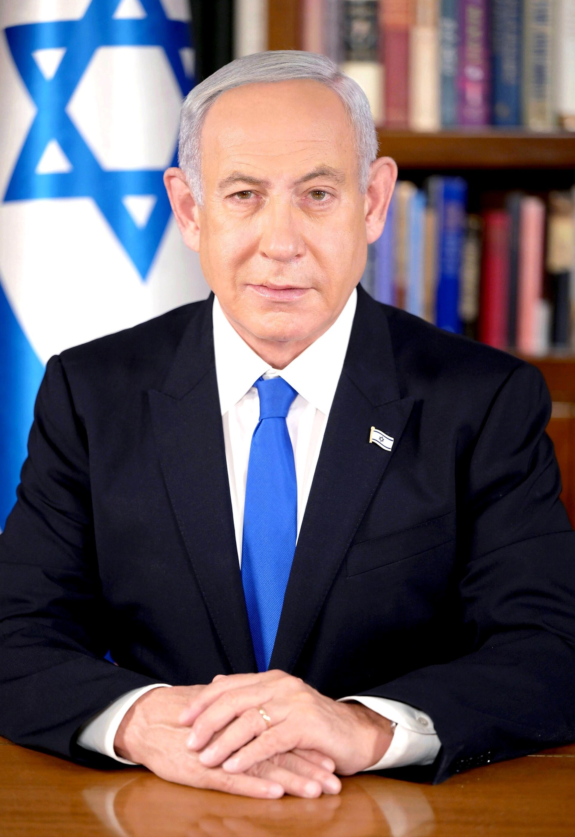 Benjamin Netanyahu in a suit sitting at a desk with his hands folded in front of him.