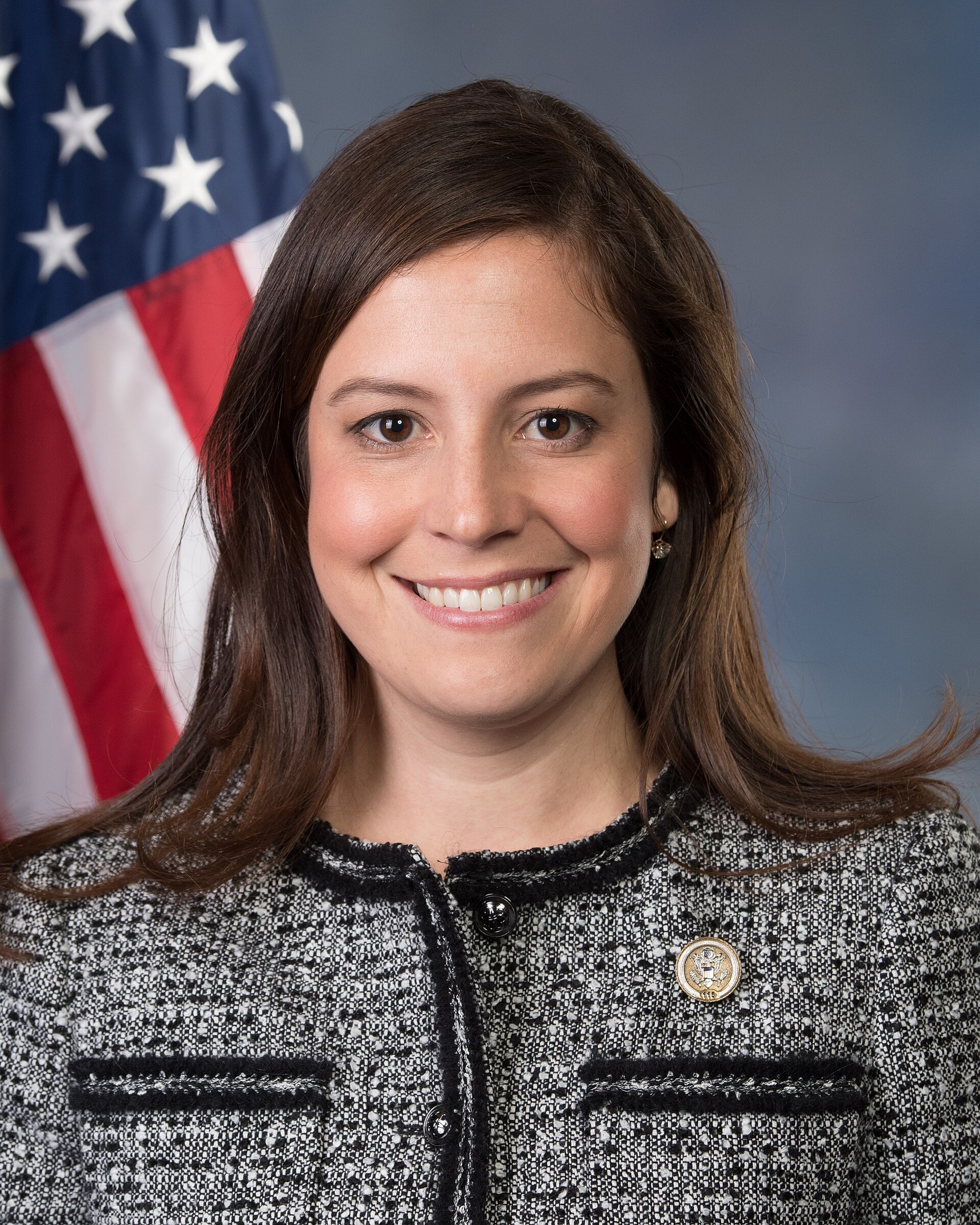 A woman wearing a black and white tweed jacket with a lapel pin stands in front of an American flag.