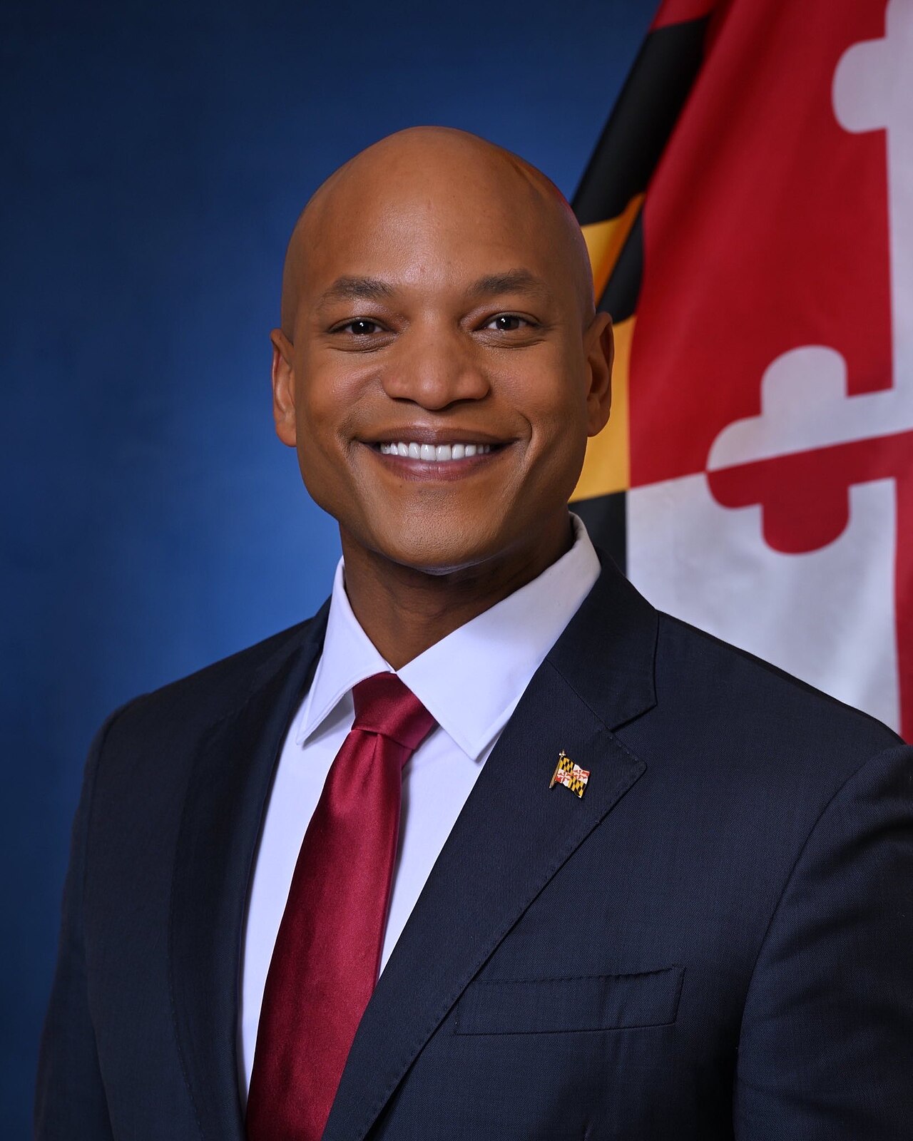 A bald man with brown skin, wearing a navy suit, white shirt, and red tie, with a Maryland flag pin on his lapel, standing in front of a Maryland state flag.