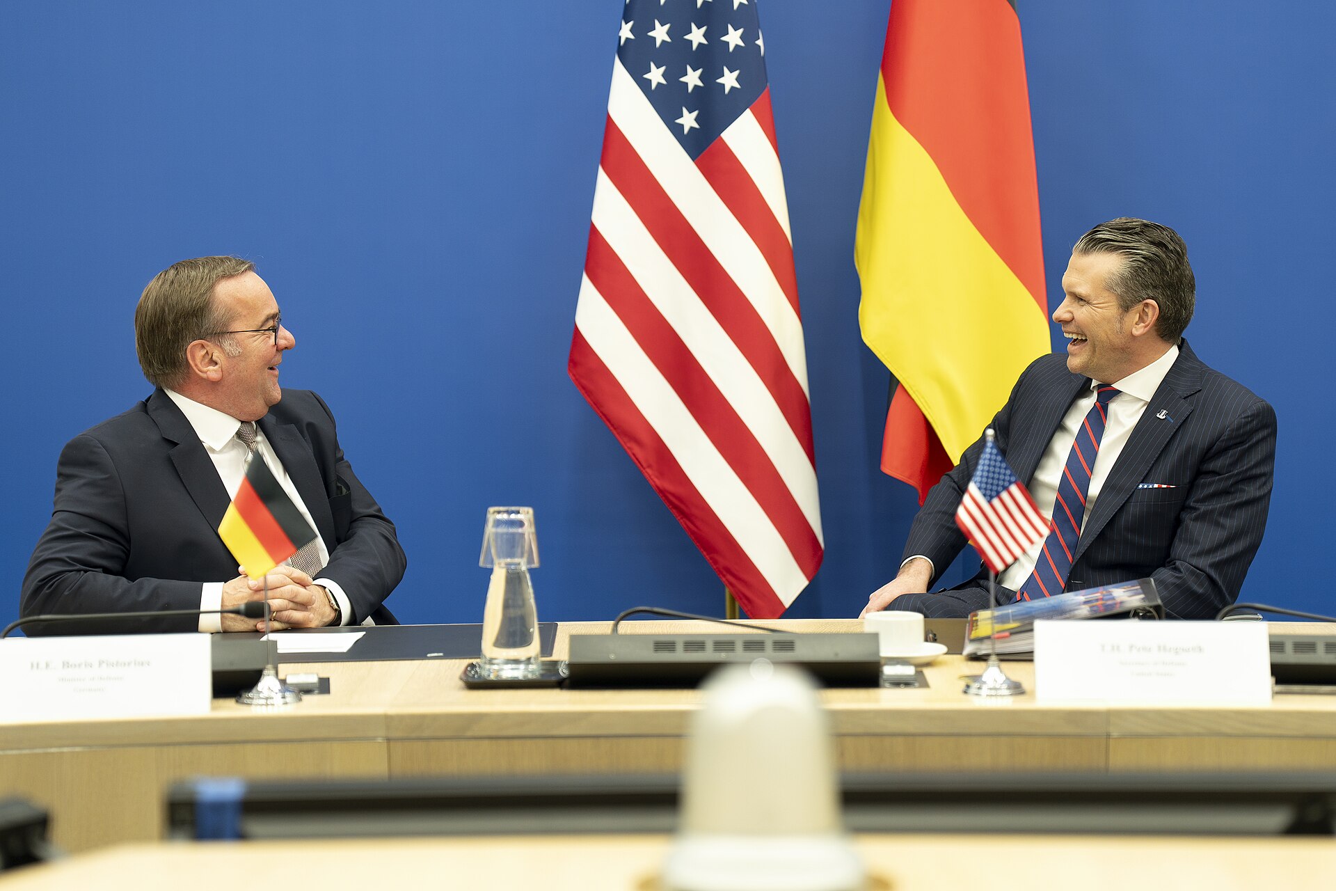 Pete Hegseth, right, in a dark suit and striped tie, sitting at a table with a name card, an American flag on the table in front of him, and an American flag hanging behind him.