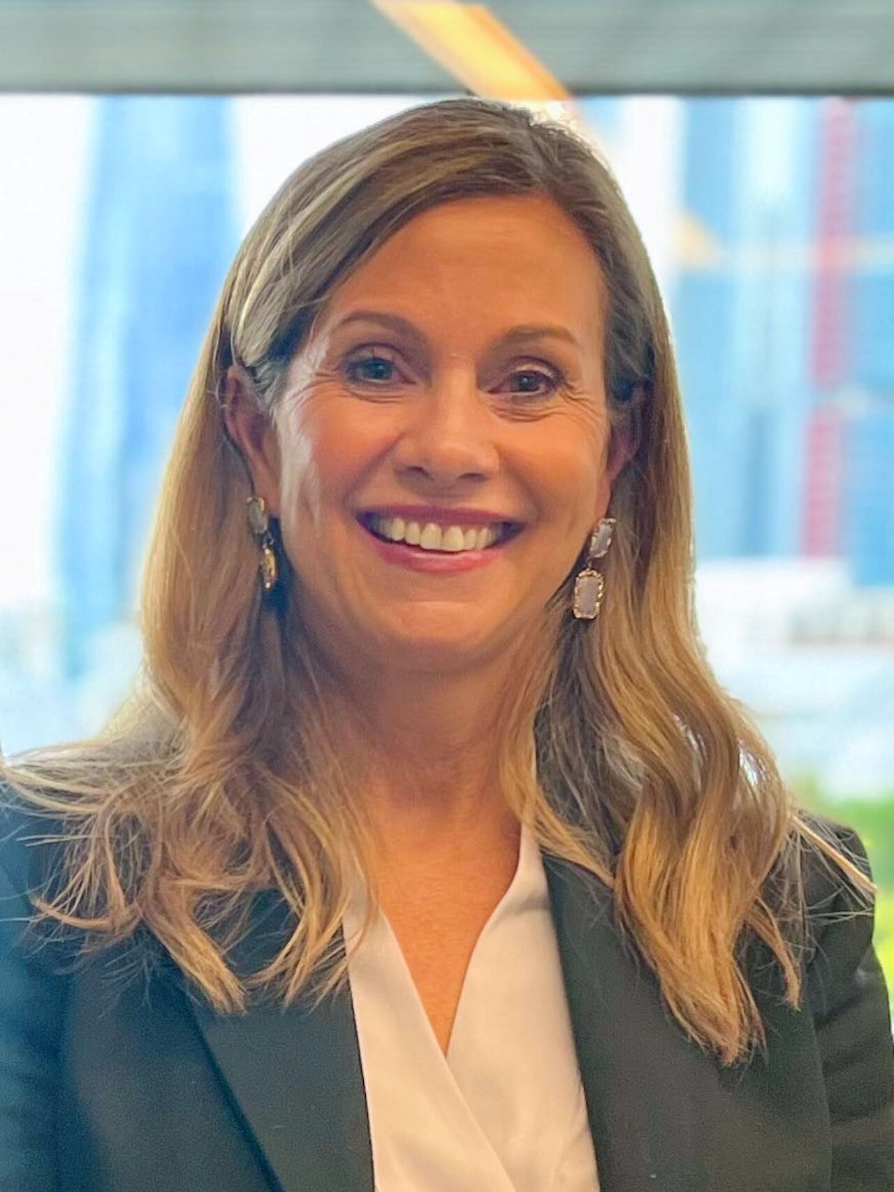 A smiling middle-aged woman with long blonde hair wearing a black blazer and white blouse.