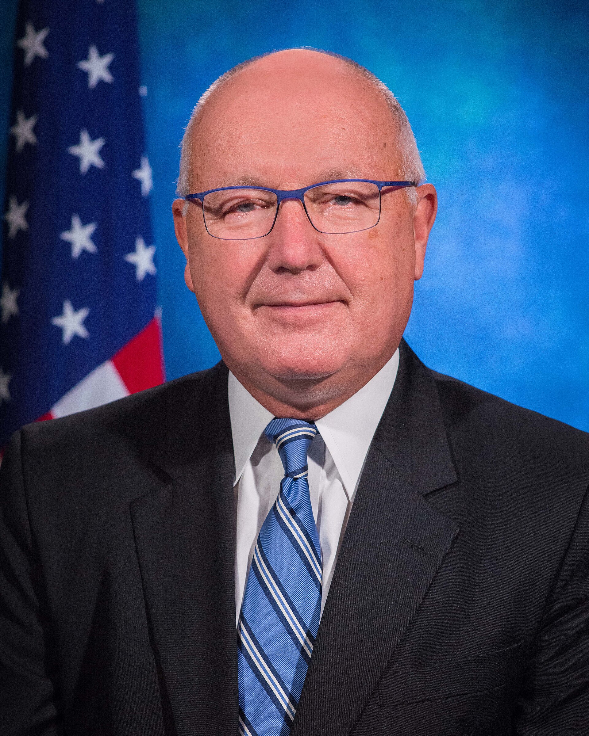 A headshot of a bald white man wearing glasses, a suit, and a blue striped tie, with the American flag and a blue background behind him.