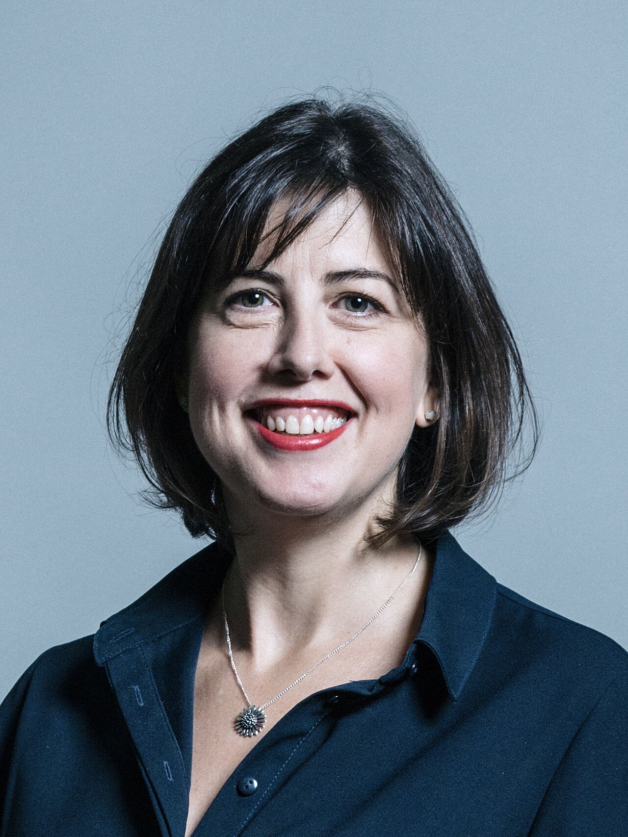 A smiling woman with short dark hair, wearing a dark blue collared shirt and a silver necklace with a pendant, against a light gray background.