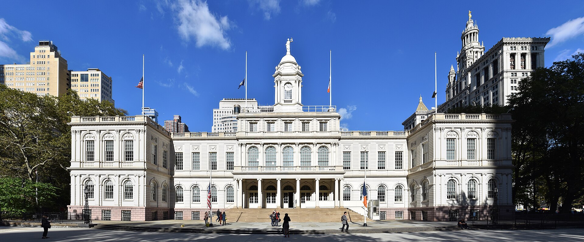 A wide shot of New York City Hall under a clear blue sky, with flags flying at half-mast.