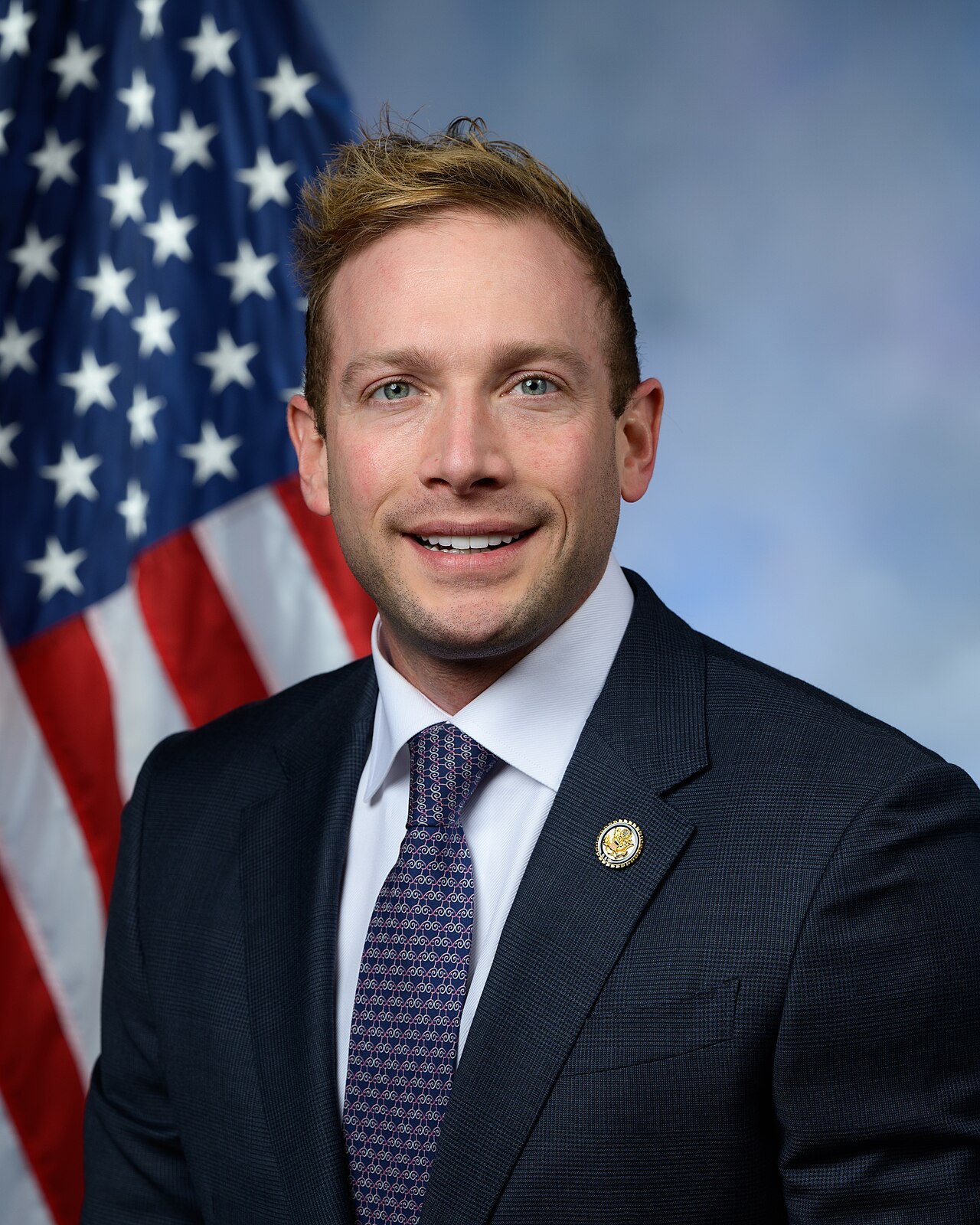 A smiling man in a suit and tie stands in front of an American flag. He has short brown hair and wears a dark suit, white shirt, and patterned tie. A pin adorns his lapel.