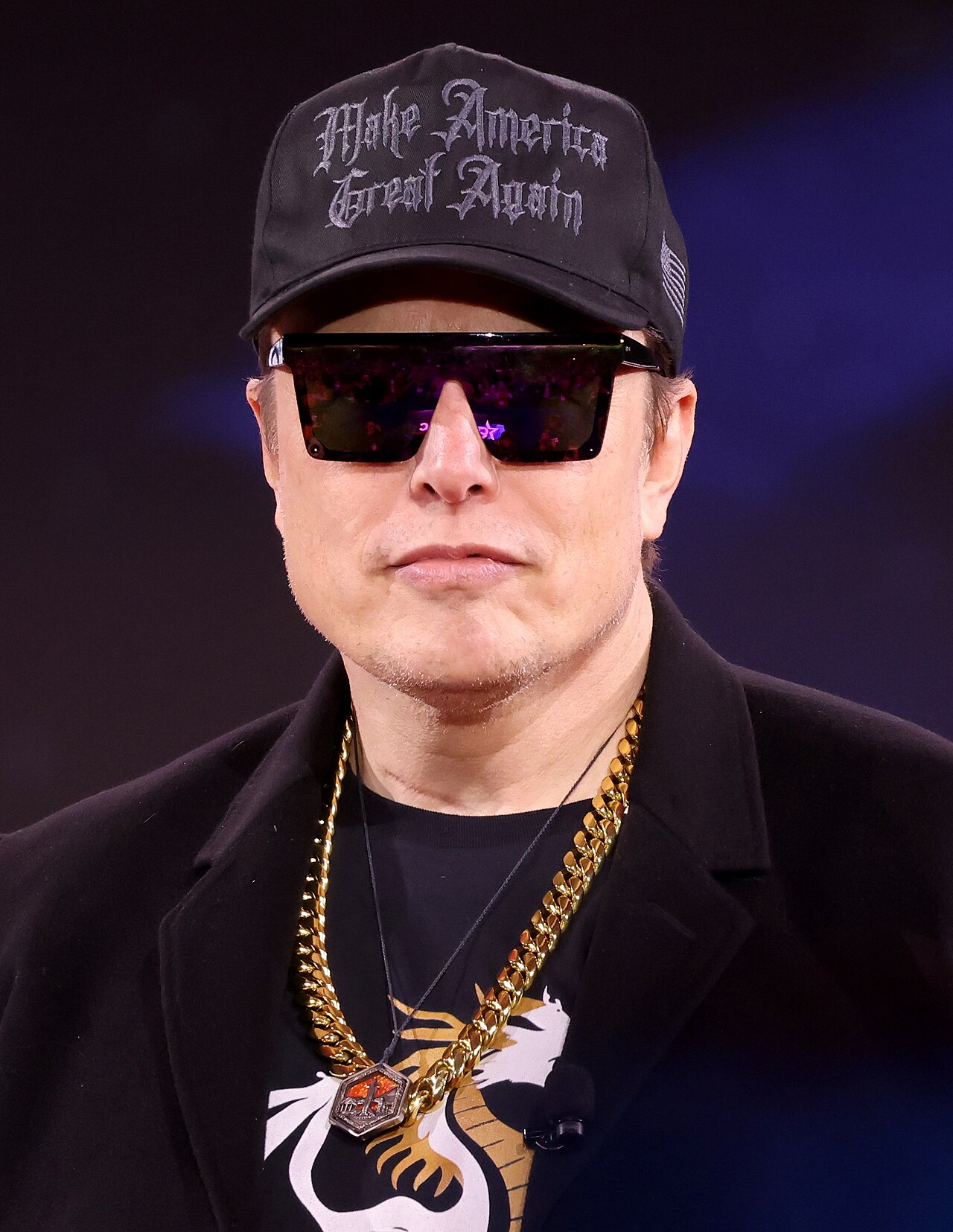 Elon Musk in a black MAGA cap, dark sunglasses, and a gold chain, at a microphone.
