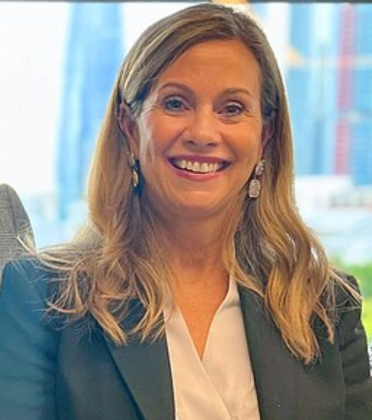 A smiling middle-aged woman with long blonde hair wearing a dark blazer and white blouse.