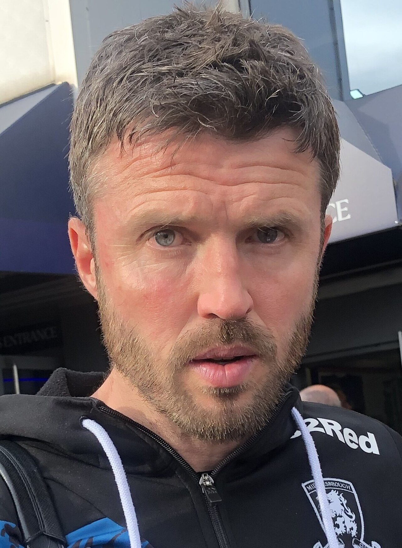 A close-up photo of a middle-aged man with short brown hair and a beard, wearing a black hoodie with white drawstrings and a logo.