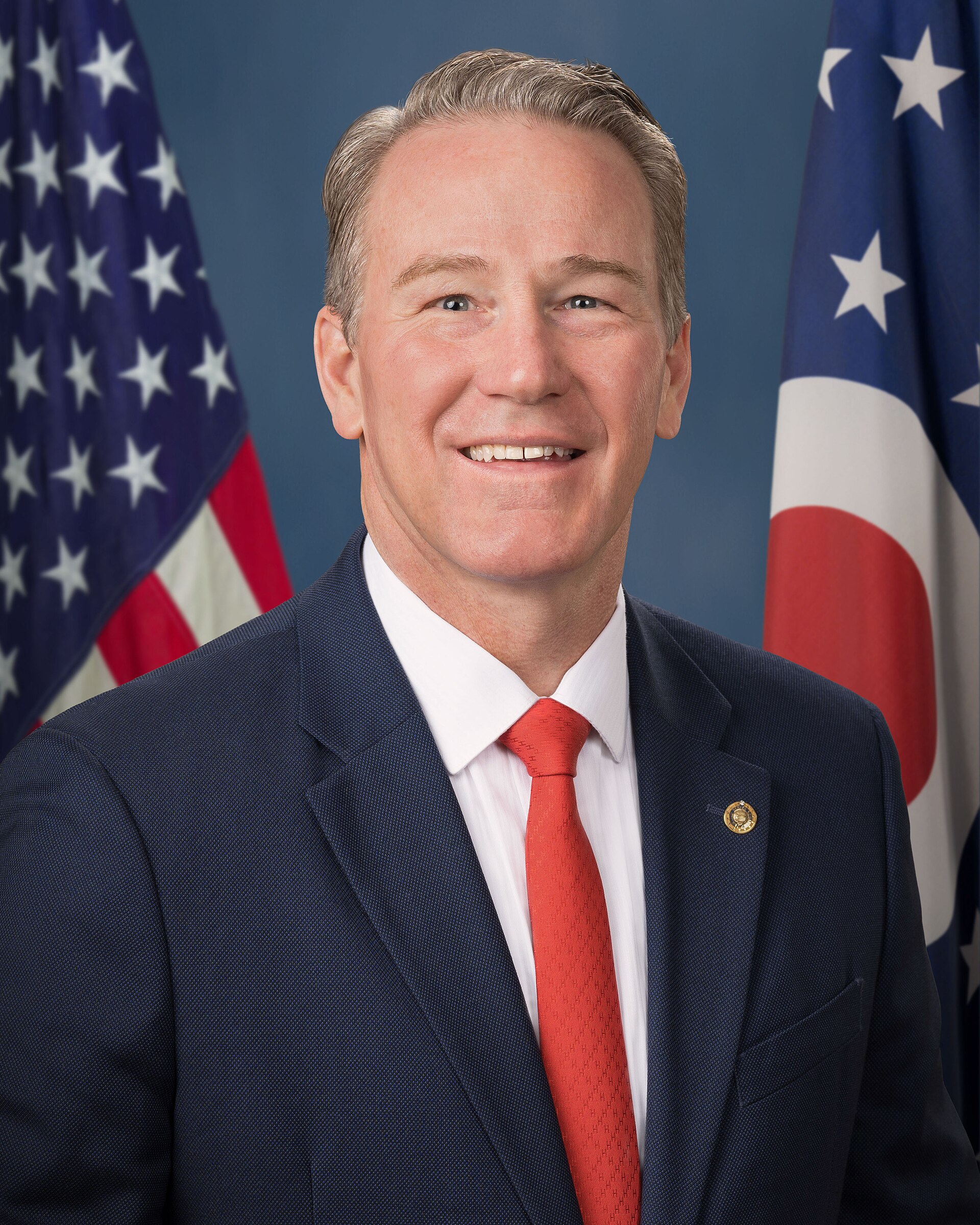 Jon Husted, smiling, wearing a blue suit and red tie, with American and Ohio flags behind him.