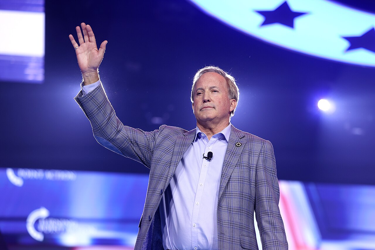 A man in a plaid suit and light blue shirt waves with his right hand raised, standing on a stage with a blue background and stars.