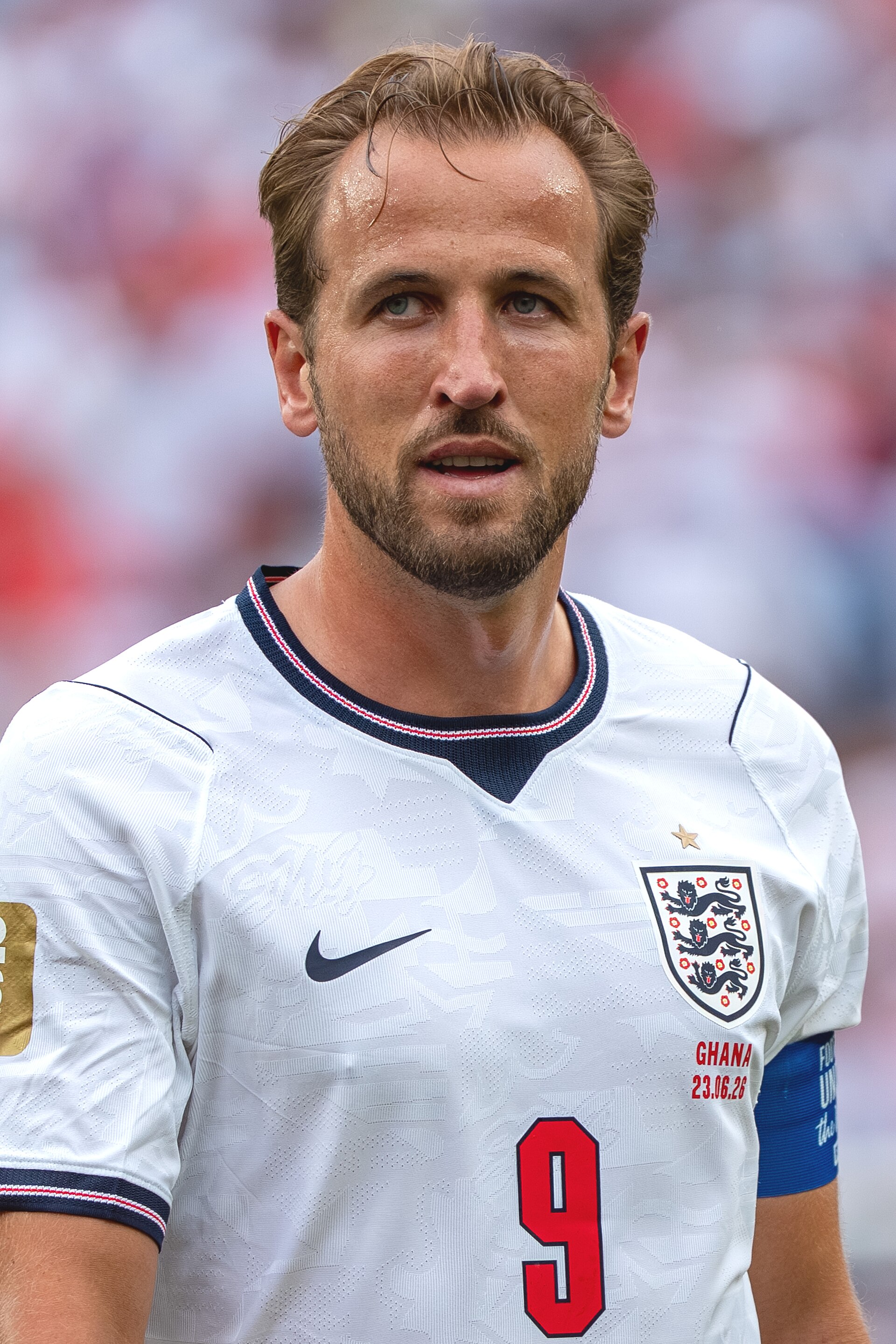 A close-up photo of a fair-skinned man with short brown hair and stubble, wearing a white England soccer jersey with a blue, red, and white collar and cuffs, a Nike check on the left chest, a Three Lions shield on the right chest, and a red number 9 on the front.