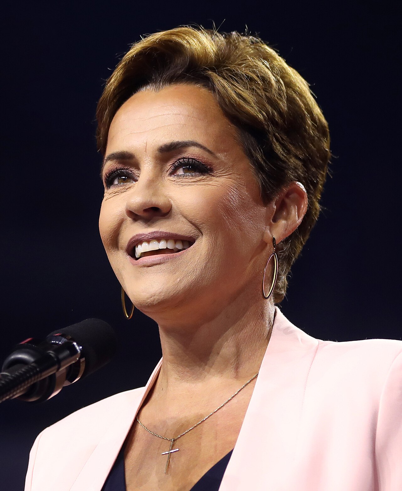 Kari Lake speaking into a microphone, smiling and wearing a pink blazer, gold hoop earrings, and a cross necklace.