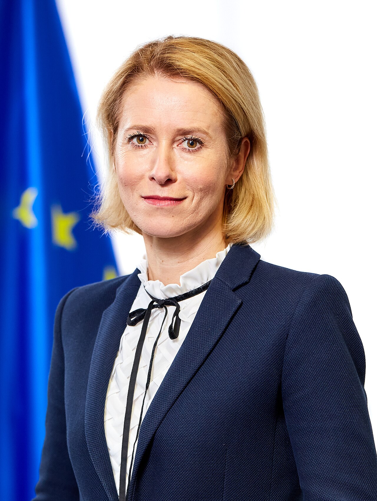 A woman with blonde hair wearing a blue blazer and white shirt, standing in front of a blue flag with yellow stars.