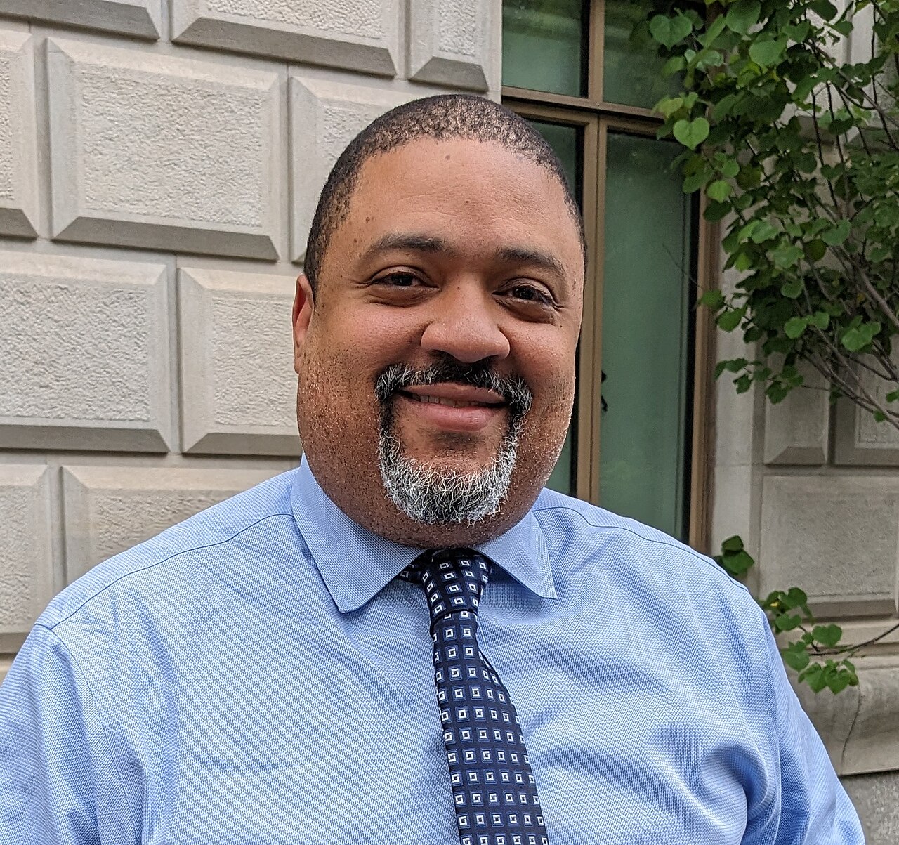 A man with short black hair and a salt-and-pepper goatee, wearing a light blue shirt and dark tie with a white square pattern, standing in front of a stone building with a window and greenery.