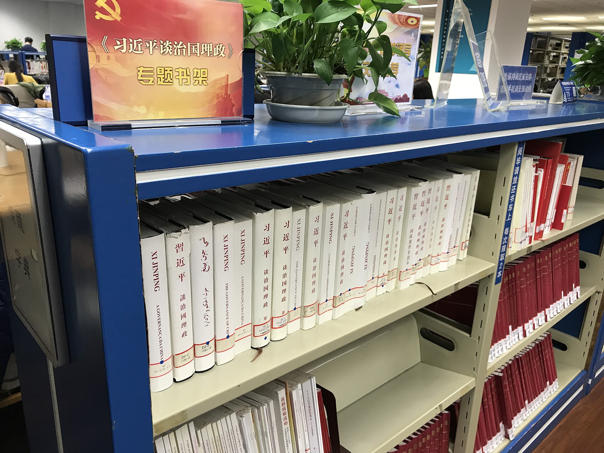 A bookshelf displaying books with "Xi Jinping" and "The Governance of China" on their spines, along with a sign featuring the Chinese Communist Party emblem and text related to Xi Jinping's governance.